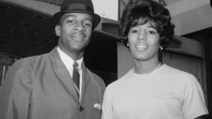 To the left an African American young man wearing a hat, suit and tie and to the right an African American young woman neatly dressed 