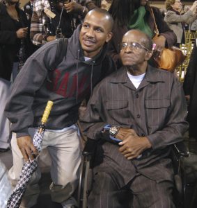 Older man seated in wheelchair on right and younger man standing to his left as they look to the left of the photo frame